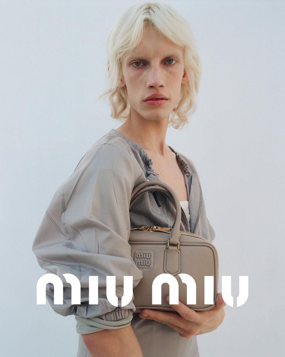 Fashion shot featuring a slender adult with blonde hair and a neutral facial expression, posing with a neutral-toned purse against a white backdrop.