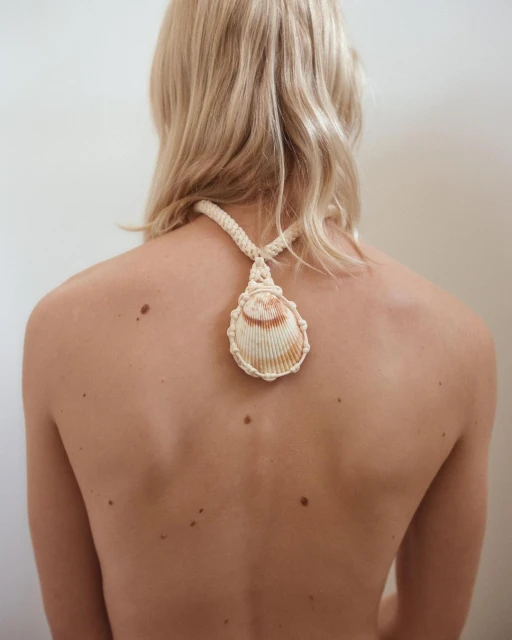 Back view of a fair-skinned person with blonde hair wearing a large seashell pendant necklace against a neutral studio backdrop.