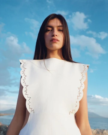 Low-angle shot of a woman with long dark hair wearing a white cutwork top against an ocean and sky background.