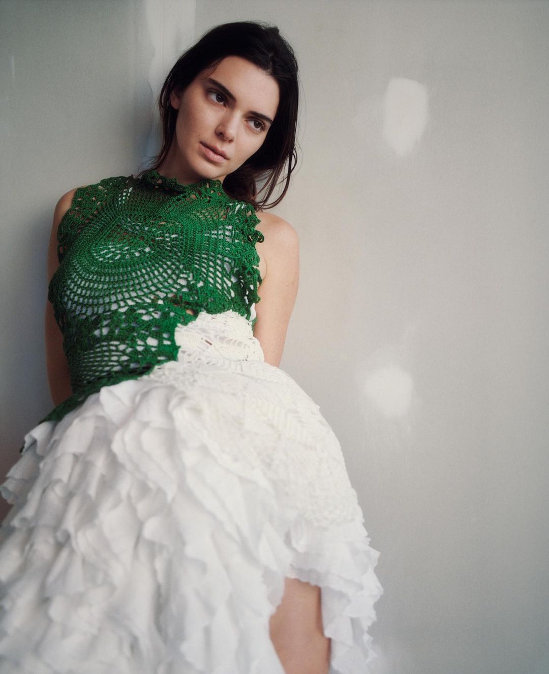 Portrait of a fashion model wearing a green and white lace dress, leaning against a grey wall, with long dark hair and neutral expression.