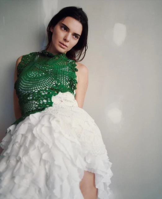 Portrait of a fashion model wearing a green and white lace dress, leaning against a grey wall, with long dark hair and neutral expression.