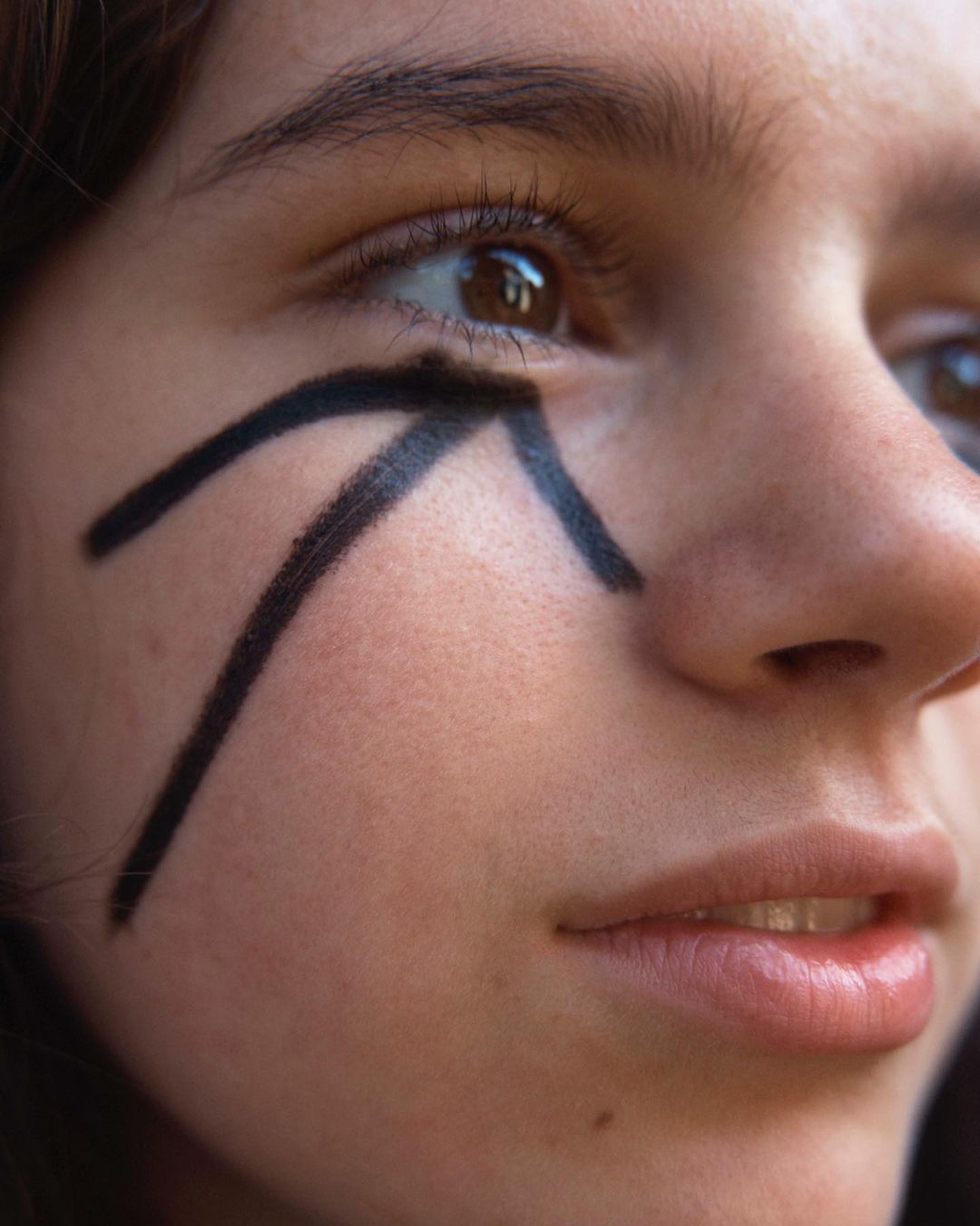 Close-up shot of a young adult woman's face with black graphic makeup around her eye, looking towards the right side of the frame.