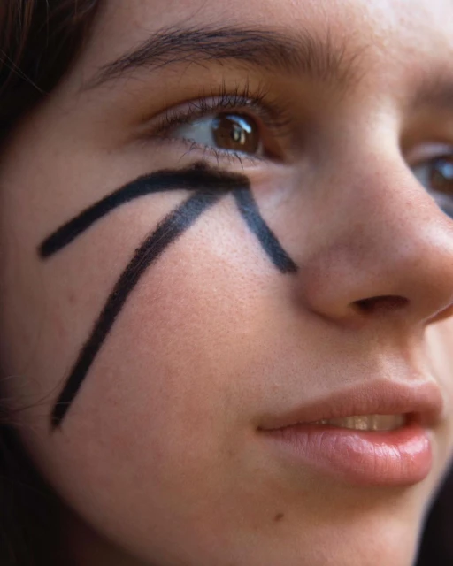 Close-up shot of a young adult woman's face with black graphic makeup around her eye, looking towards the right side of the frame.