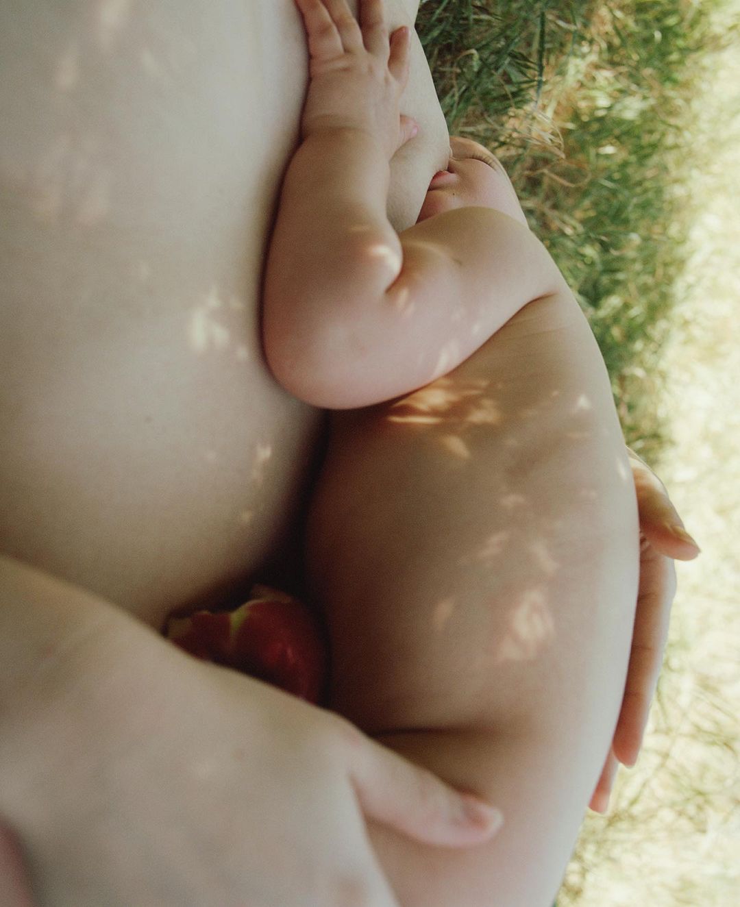 Close-up view of a baby cradled in someone's arms outside, with dappled light creating shadows; an apple sits between them.