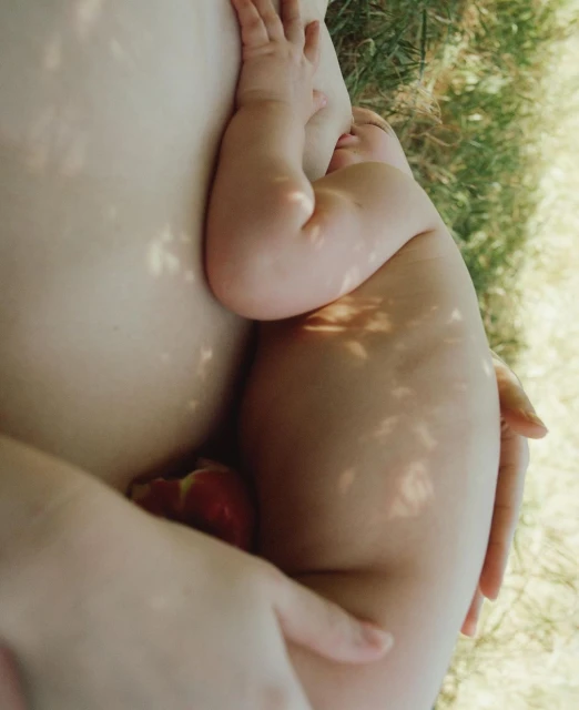 Close-up view of a baby cradled in someone's arms outside, with dappled light creating shadows; an apple sits between them.
