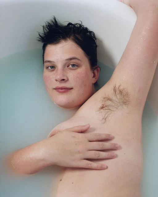 Portrait of a woman in a bathtub filled with milky water, looking at the camera with a slight smile and relaxed pose. Arm raised.