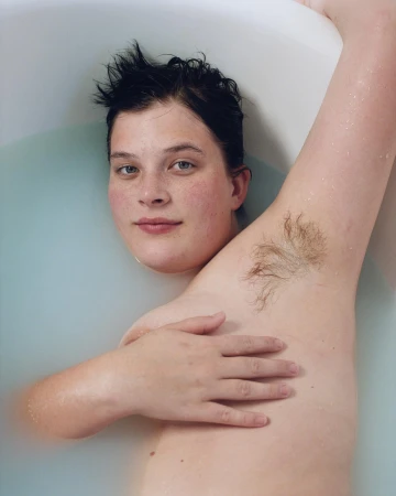 Portrait of a woman in a bathtub filled with milky water, looking at the camera with a slight smile and relaxed pose. Arm raised.