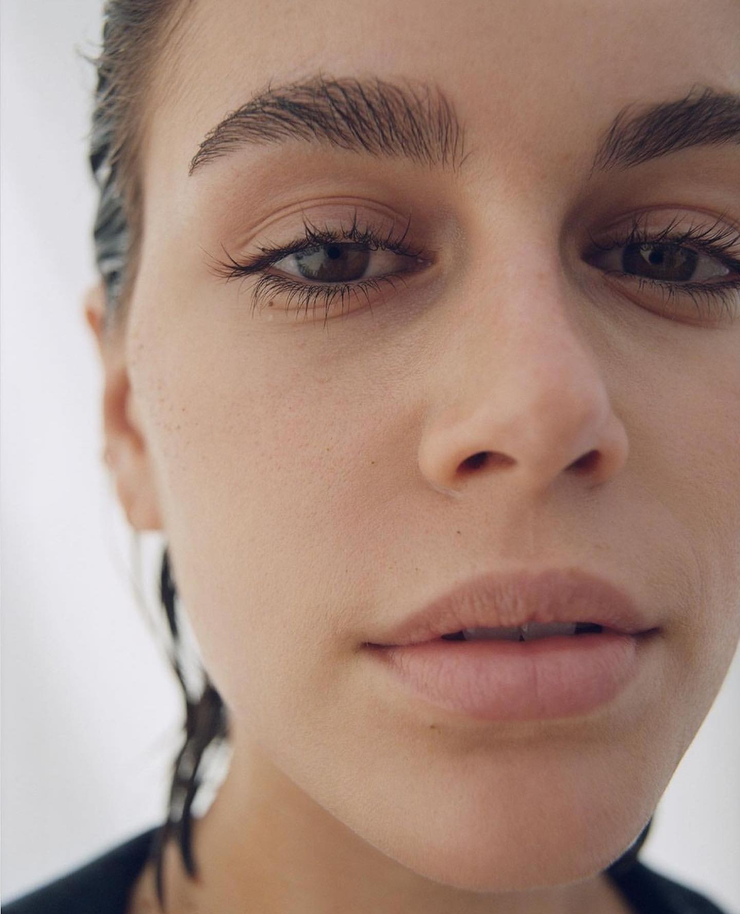 Close-up portrait of a woman with fair skin, brown eyes, and dark hair pulled back; gazing directly forward with a neutral expression against a white background.