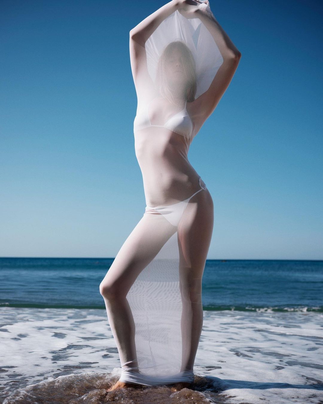 Woman in white swimsuit and sheer fabric poses on a beach with blue sky and ocean backdrop, creating a light and airy feel.