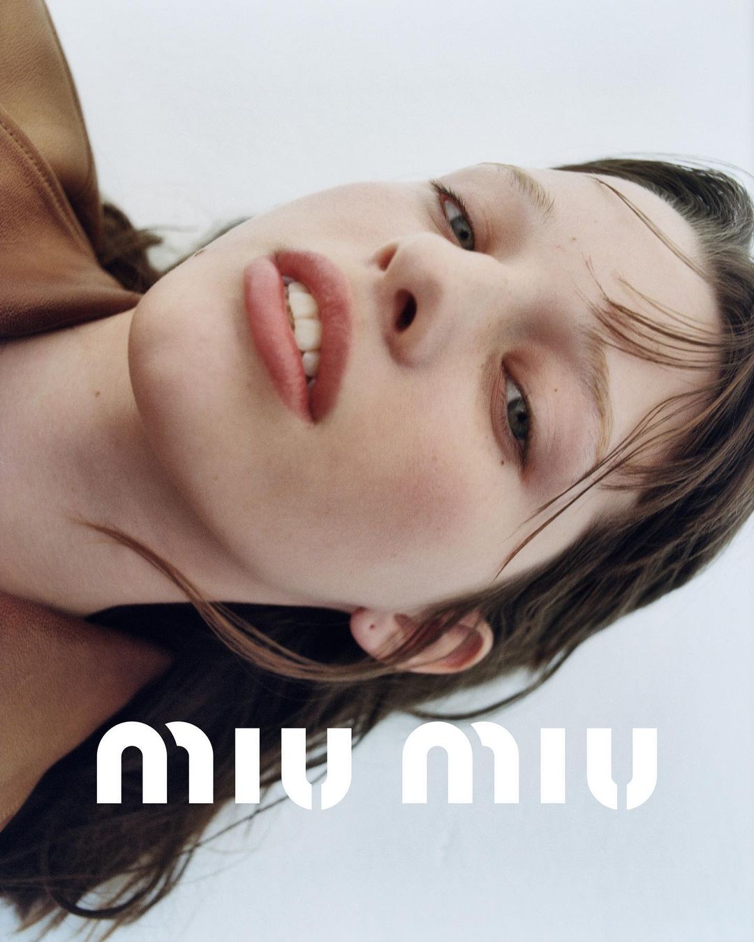 Close-up studio portrait of a young woman with fair skin and brown hair, wearing a brown leather jacket and Miu Miu branding visible.