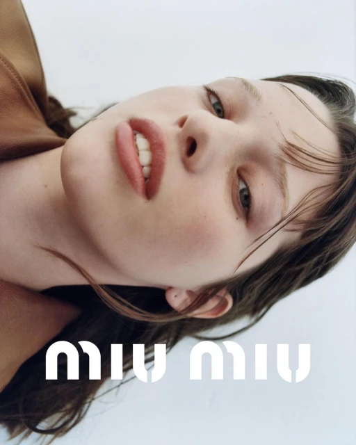 Close-up studio portrait of a young woman with fair skin and brown hair, wearing a brown leather jacket and Miu Miu branding visible.