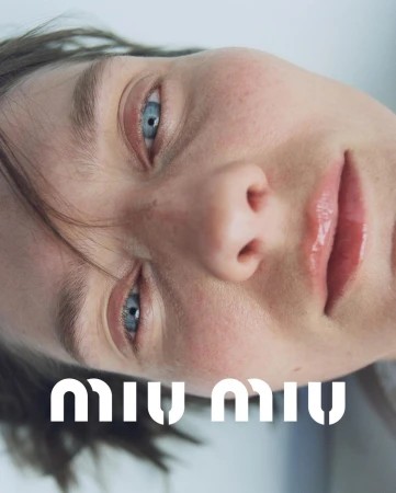 Close-up portrait of a fair-skinned adult female with striking blue eyes and glossy lips, featuring the 'Miu Miu' logo at the bottom.
