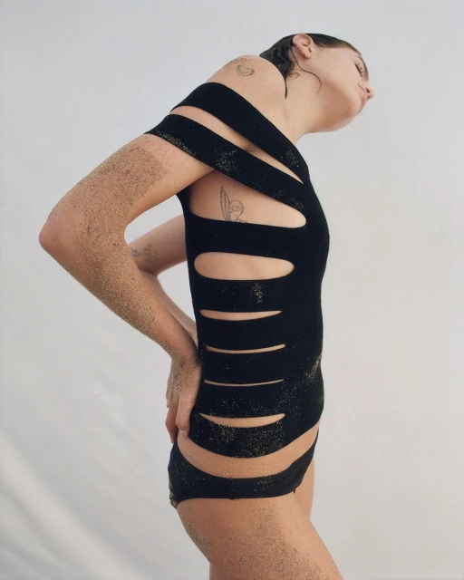 A woman poses in a black cutout bodysuit with sand on her skin against a white background, looking up with a neutral facial expression.