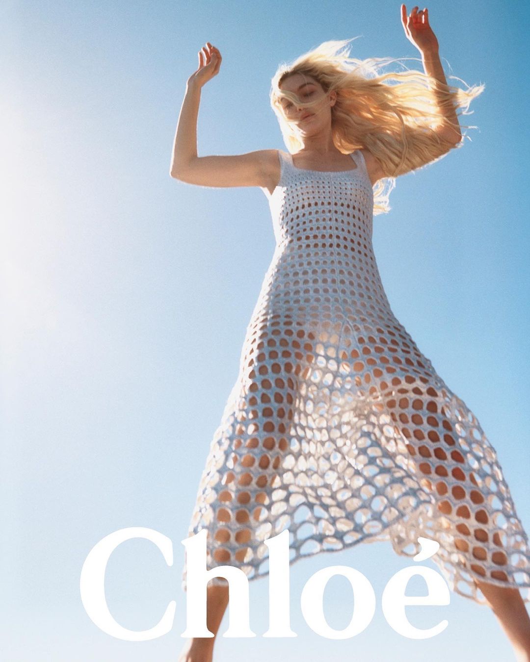 Fashion shot of a blonde woman in a white mesh dress, arms raised, set against a bright blue sky, promoting Chloe fashion brand.