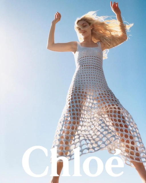 Fashion shot of a blonde woman in a white mesh dress, arms raised, set against a bright blue sky, promoting Chloe fashion brand.