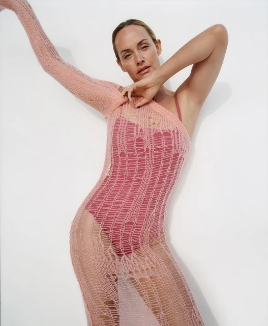 A woman poses in a studio wearing a red one-piece swimsuit and a long, pink, loosely knit sheer dress with a one-shoulder sleeve.