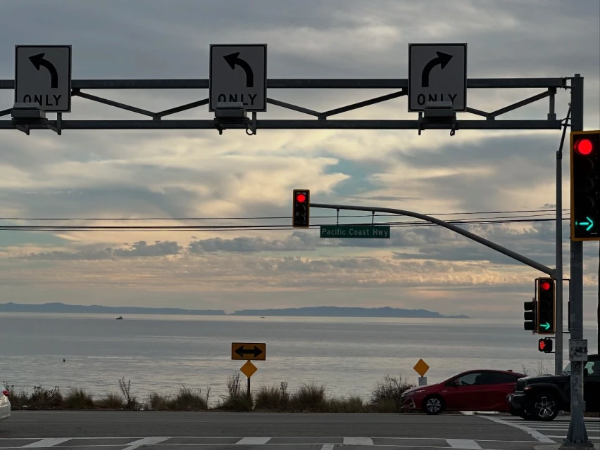 A coastal highway intersection with traffic lights and directional signs, with a cloudy sky and ocean visible in the background.