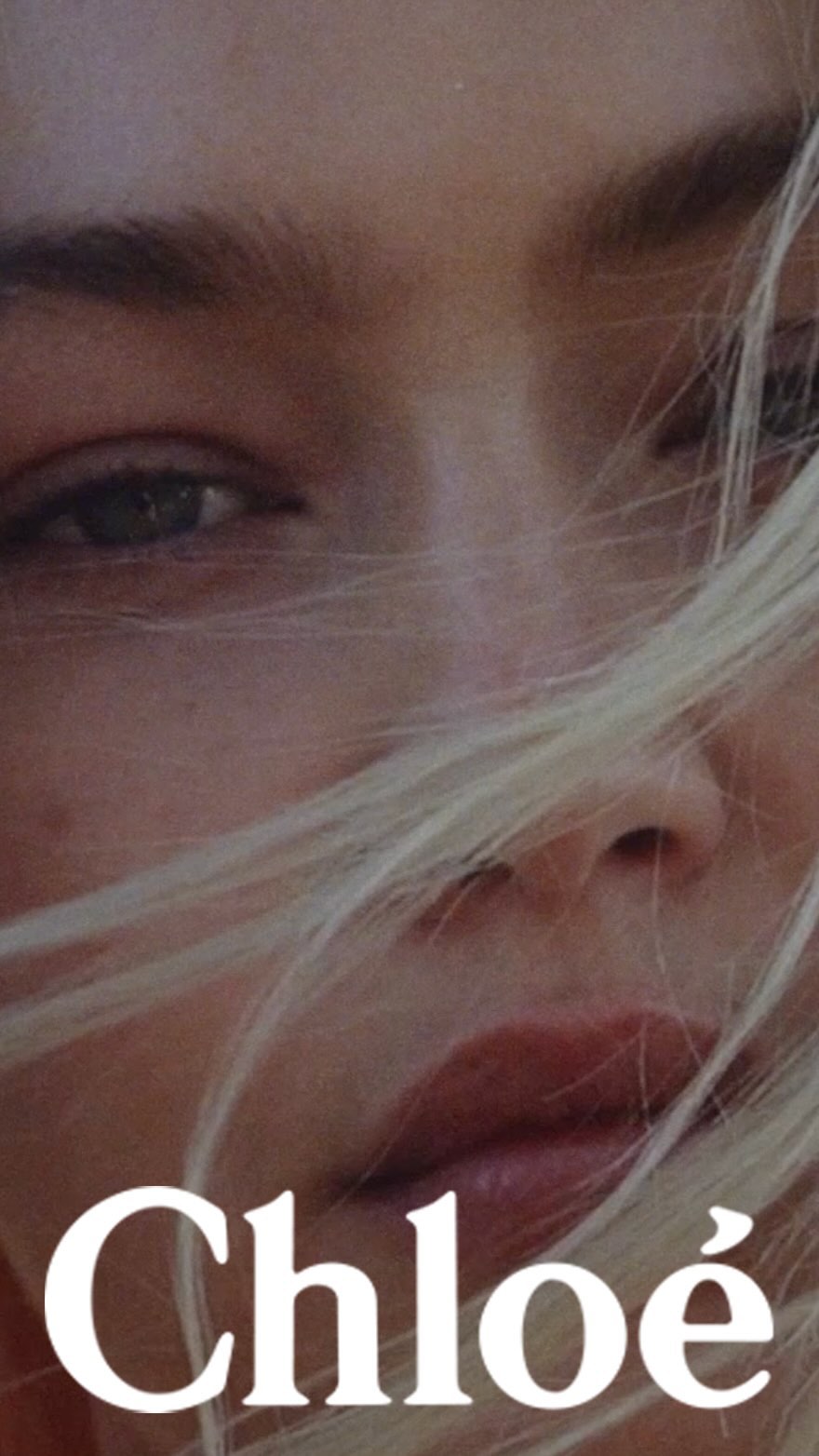 Close-up portrait of a woman with blonde hair blowing across her face and white Chloe text overlaid at the bottom.