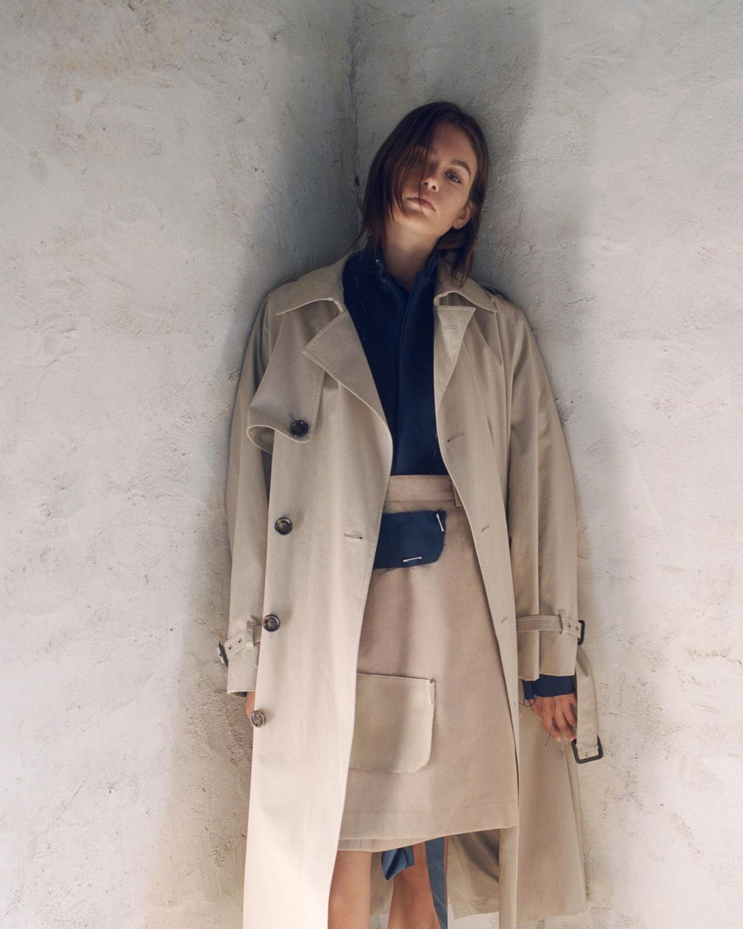 Fashionable woman in studio wearing tan trench coat and skirt, posing against a neutral wall. Shoulder-length dark hair, serious expression.