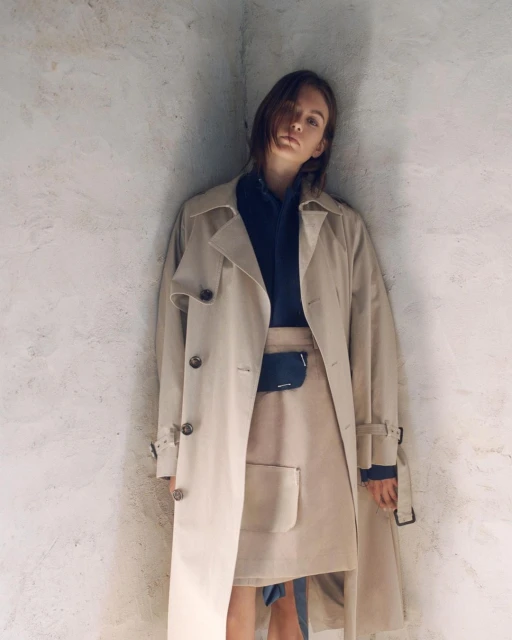Fashionable woman in studio wearing tan trench coat and skirt, posing against a neutral wall. Shoulder-length dark hair, serious expression.