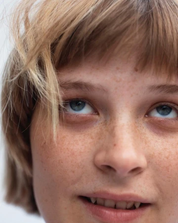 Close-up portrait of a fair-skinned young adult with blonde hair and freckles looking up with a slight smile on a neutral background.