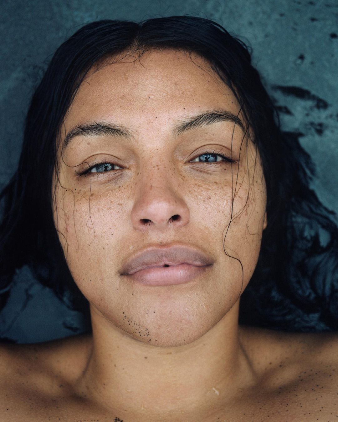 Close-up portrait of a young woman with dark hair and freckles, her face partially submerged in water, creating a natural, serene effect.