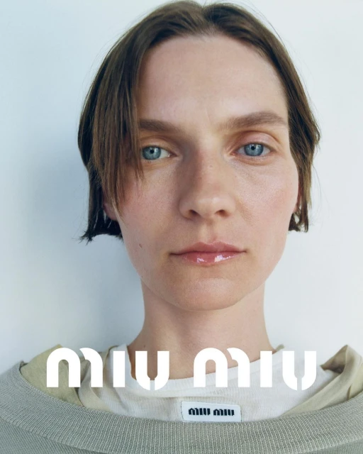 Close-up studio shot of a model with short brown hair and blue eyes, wearing a beige sweater, with the Miu Miu logo at the bottom.