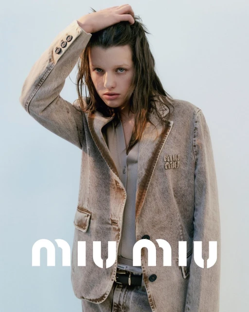 A young woman poses in a fashionable denim jacket with the Miu Miu logo against a solid backdrop, creating a high-fashion portrait.