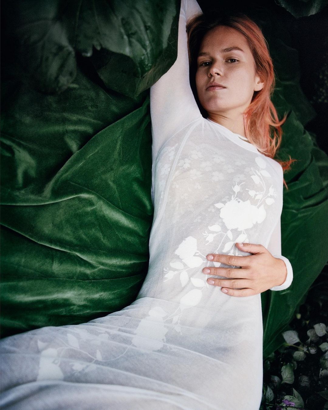 Woman with pink hair in white dress reclines in botanical setting with large green leaves. Fashion and beauty portrait in nature.