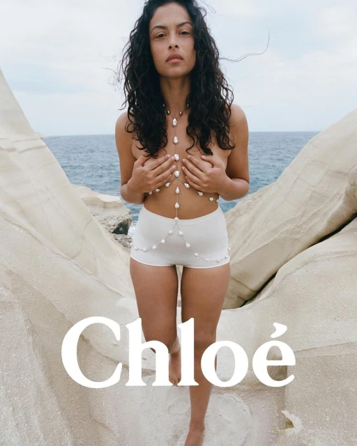 A young woman poses on a rocky beach wearing a pearl body chain and knit shorts, in a fashion-style shot against a blue sea.