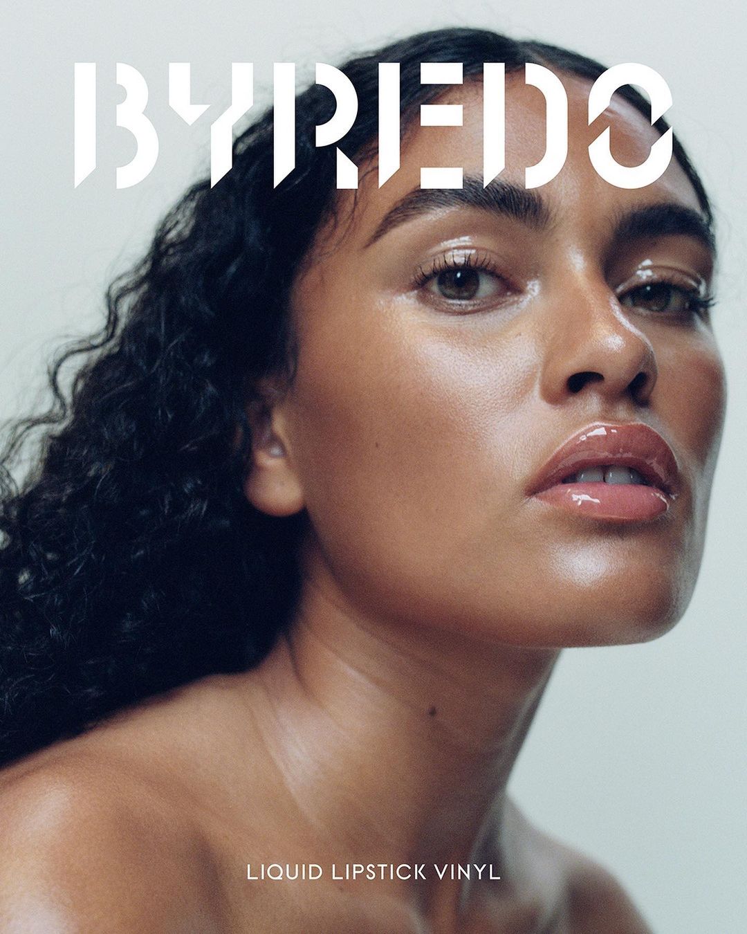 Close-up portrait of a woman with dark curly hair and glossy lips in a beauty advertisement. The image features 'BYREDO' in bold text.