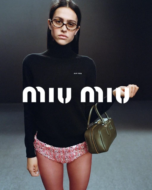 A model poses indoors wearing a black turtleneck, sequined shorts, and glasses, holding a small olive-green handbag, with the MIU MIU logo.