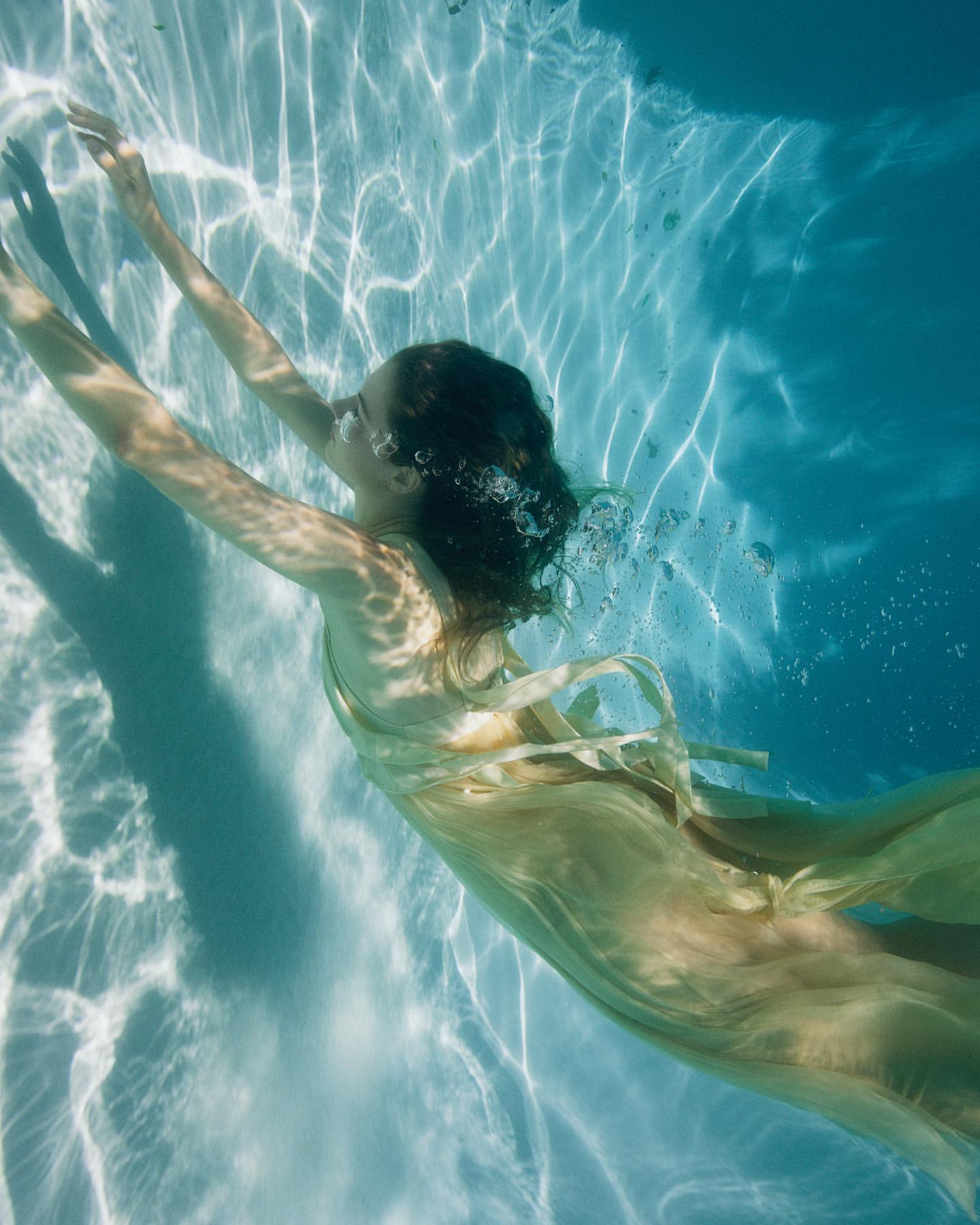 Underwater shot of a woman in a flowing yellow dress swimming in a pool, surrounded by sunlight patterns on the water's surface, serene and ethereal.