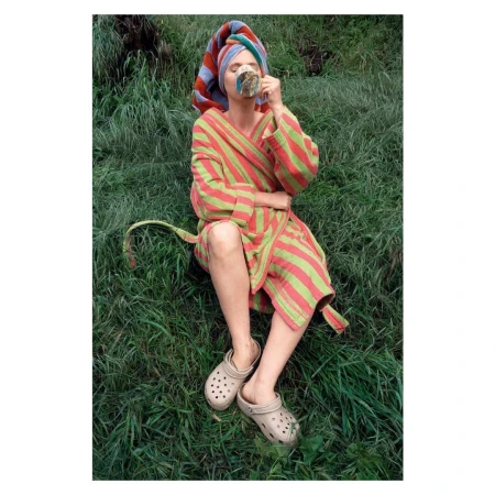 Woman in striped robe and towel turban sitting in green grass, drinking from a mug, wearing Crocs, enjoying a refreshing outdoor moment.