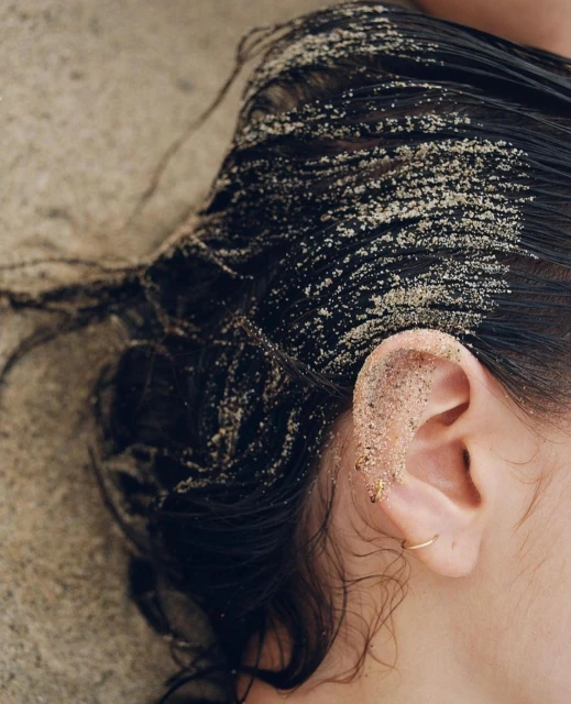 Close-up shot of a person's ear and hair covered in sand against a sandy background. The individual is wearing a gold hoop earring.