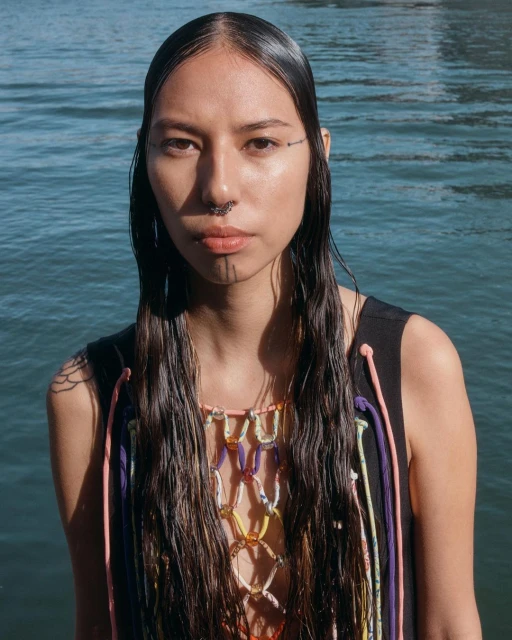 Portrait of a woman with long dark hair, face tattoos, and beaded top, standing outdoors with rippling blue water in the background.