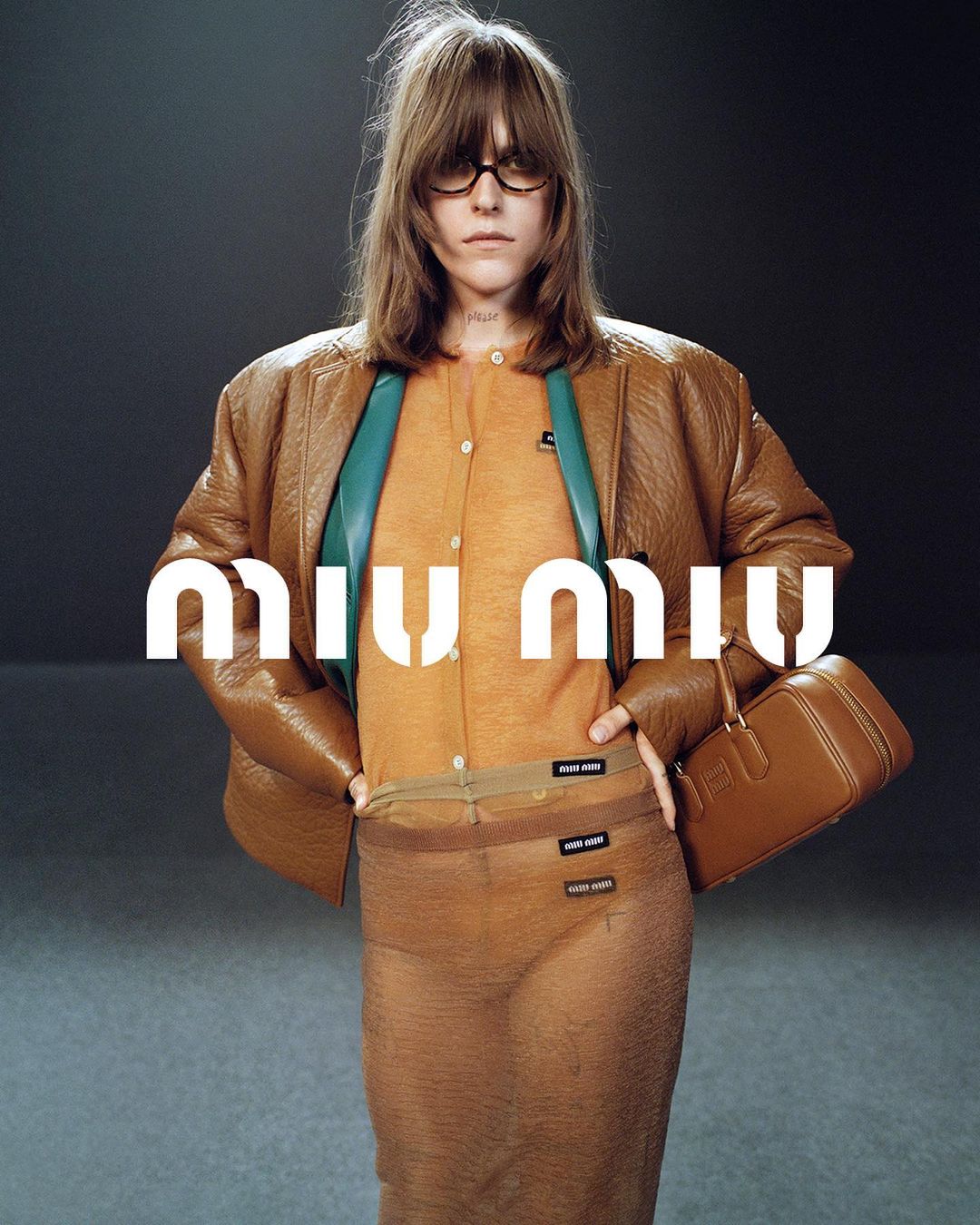 Fashion image features a woman in a sheer brown skirt and orange top, wearing glasses and a tan jacket, posed against a grey background.