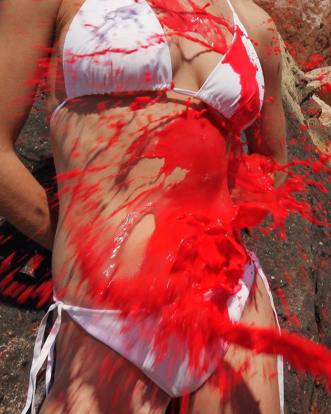 Partial view of a woman in a bikini covered in vibrant red paint, artistic and eye-catching, against a natural rock background.