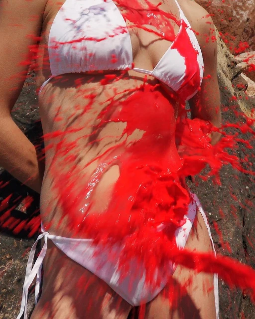 Partial view of a woman in a bikini covered in vibrant red paint, artistic and eye-catching, against a natural rock background.