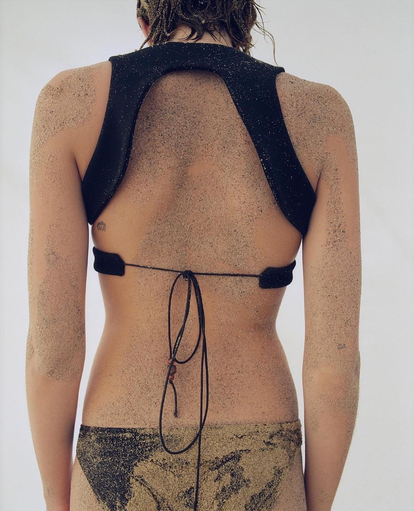 Back view of a female model wearing a black top and bikini bottom covered in sand, posing in a bright indoor photo studio.