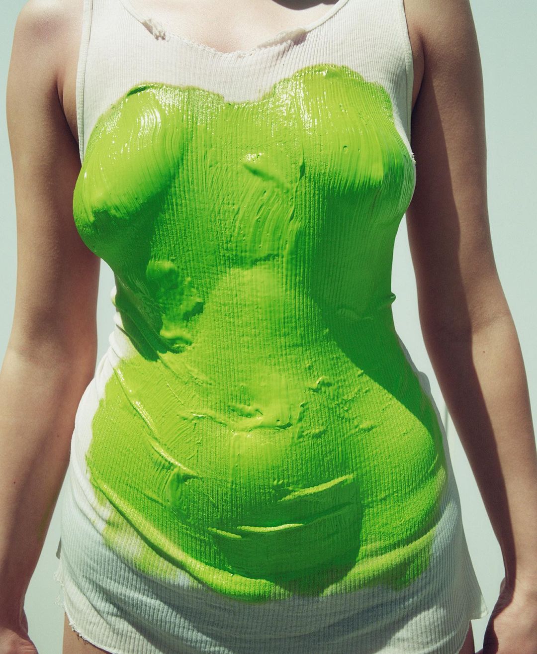 Torso of a person covered in green paint, wearing a white ribbed tank top, set against a bright white background. Conceptual art.