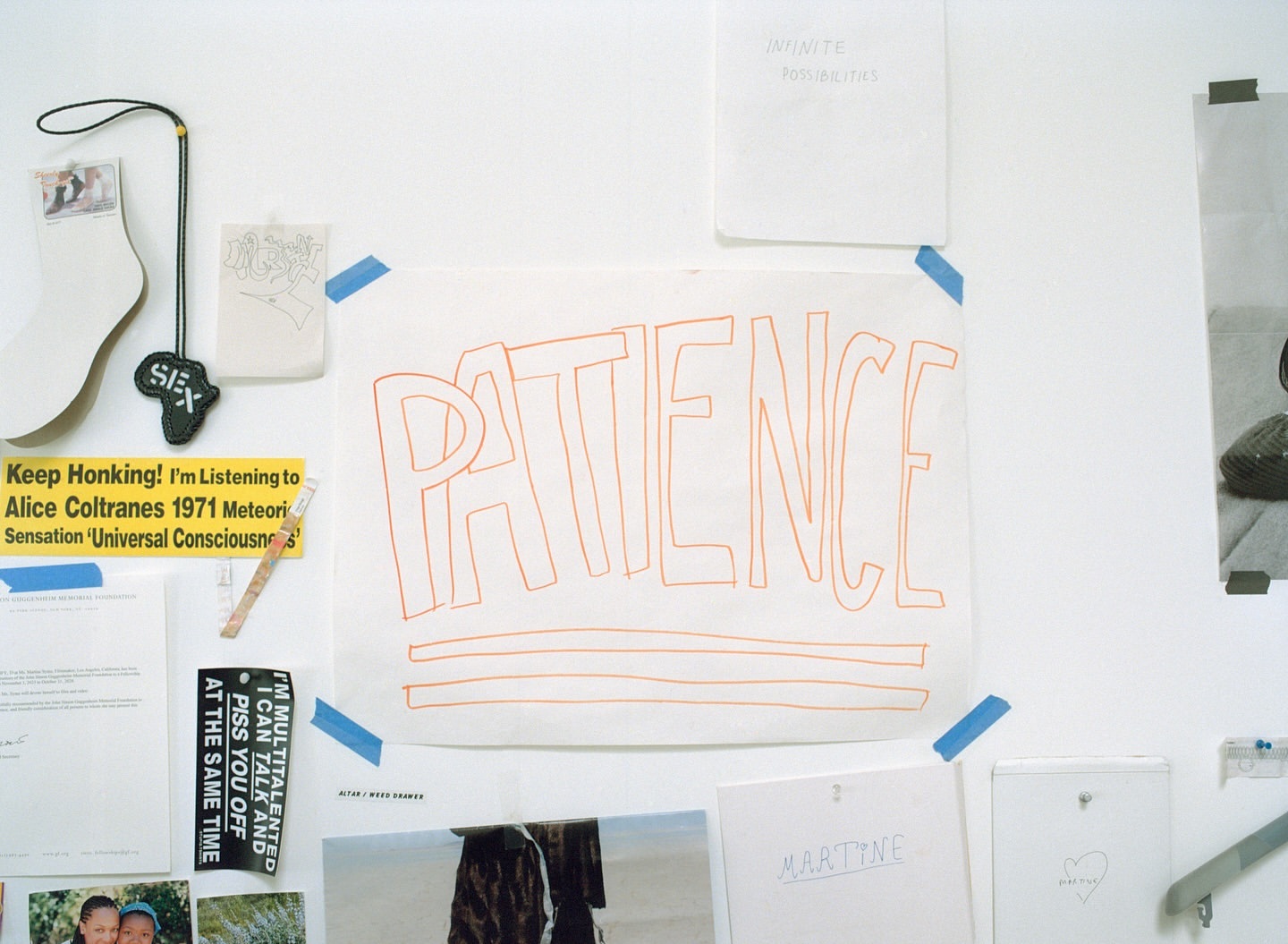 White wall with various papers, notes, and drawings attached with tape. The word 'Patience' is prominently displayed on one sheet.