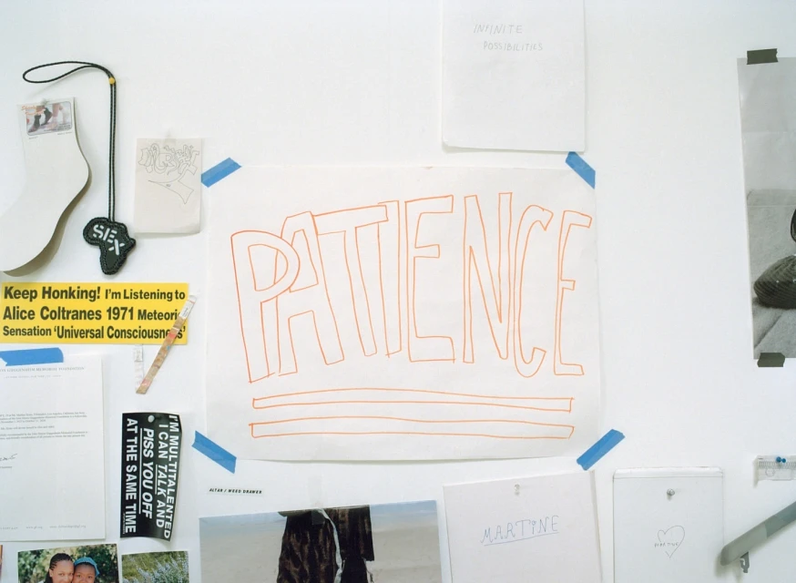 White wall with various papers, notes, and drawings attached with tape. The word 'Patience' is prominently displayed on one sheet.