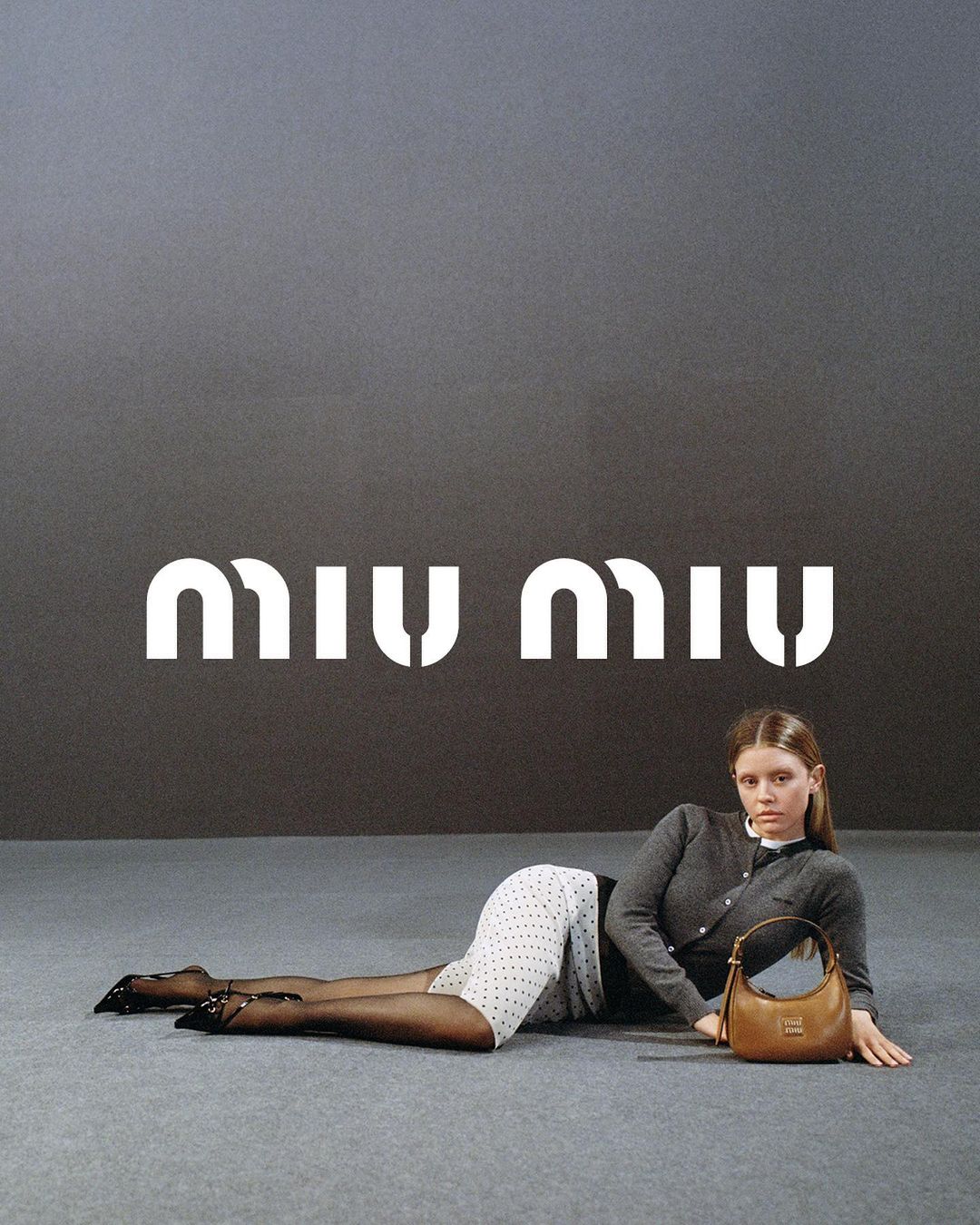 Fashion model poses with Miu Miu handbag in studio. Woman wears cardigan, polka dot skirt, and black tights against gray background.