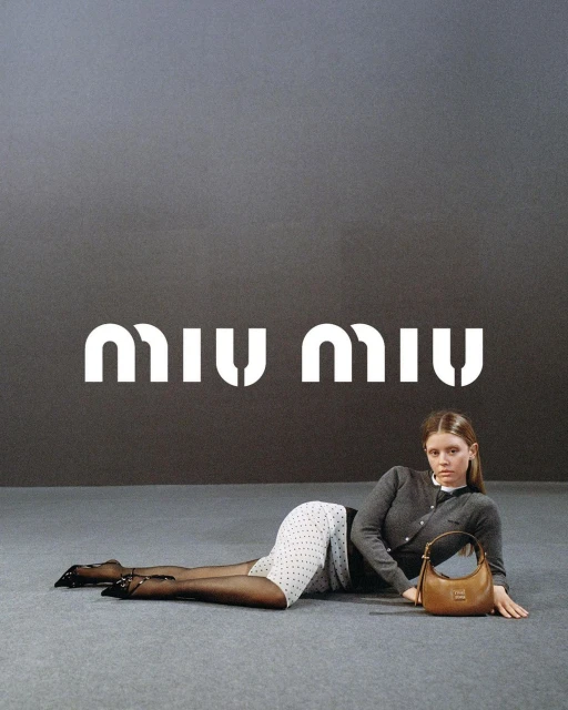 Fashion model poses with Miu Miu handbag in studio. Woman wears cardigan, polka dot skirt, and black tights against gray background.
