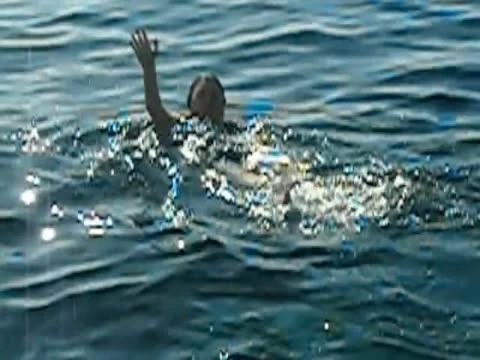 Person swimming in deep blue water with arm raised, seemingly waving or calling for help, under natural light in an ocean environment.