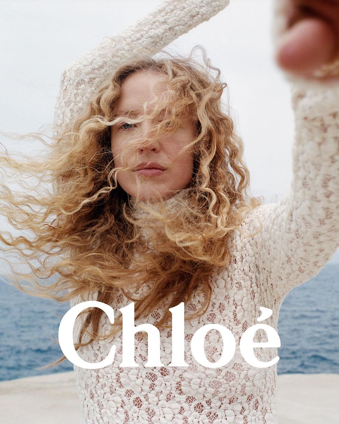 Woman with long blonde curly hair wearing a white textured lace turtleneck sweater with arms raised against an ocean background