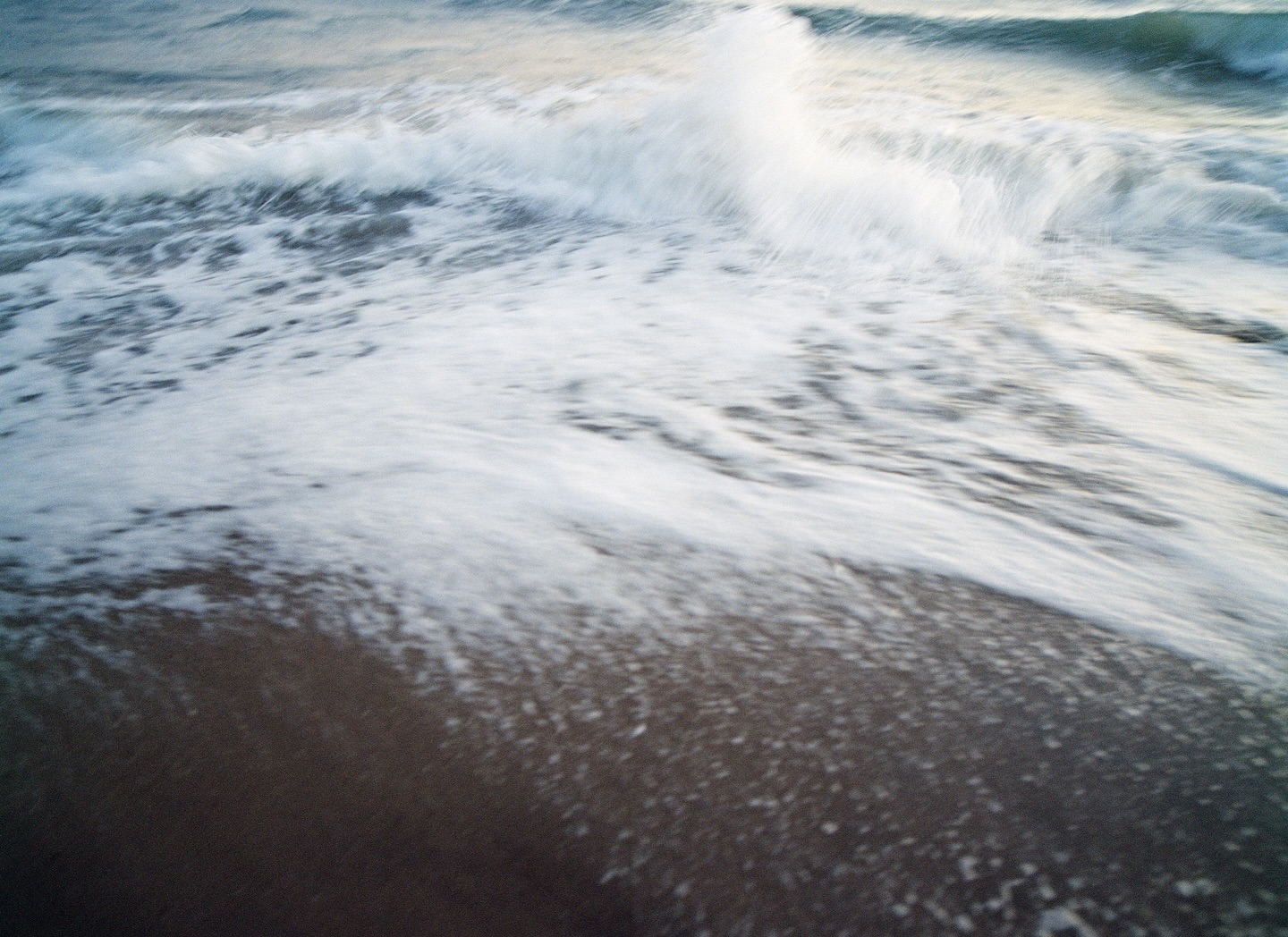 Seascape of waves crashing on a sandy beach with ocean foam in a blurred motion effect under soft, overcast, natural lighting.