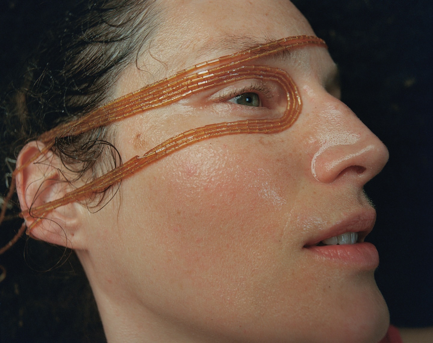 Close-up of woman's face with orange beaded strands wrapped around her head, side profile against a dark background.