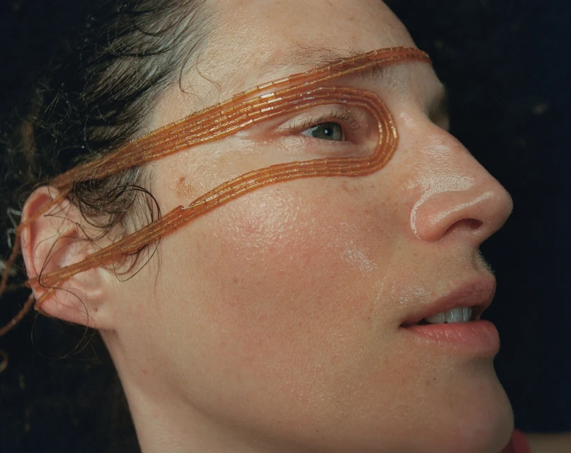 Close-up of woman's face with orange beaded strands wrapped around her head, side profile against a dark background.